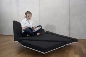 Cay Sofa with different geometric cushions turned in different directions and covered with dark grey fabric and a white geometric frame, with a person sitting on it wearing headphones and reading a magazine