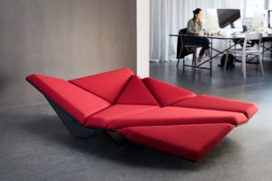Cay Sofa with different geometric cushions turned in different directions and covered with red fabric and a dark frame in a large office hall