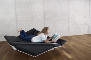 Cay Sofa with different geometric cushions turned in different directions and covered with dark grey fabric and a white geometric frame, with a person lying on it and reading a magazine