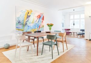 Marlon Dining Table in simple modern shape made of wood with chairs in different colors around in bright room with colorful painting on the wall