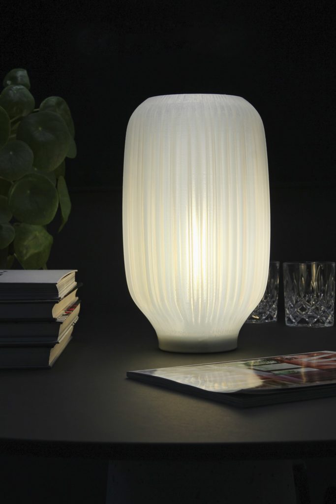 Lucia Lamp table lamp with organically folded lampshade on dark table with plant, books and glasses in background